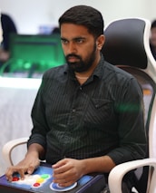 A person is seated in a chair, focused intently on something, with hands positioned on a gaming console or arcade controller. The setup appears to be indoors, with visible screens and lighting in the background, suggesting an electronic gaming environment.